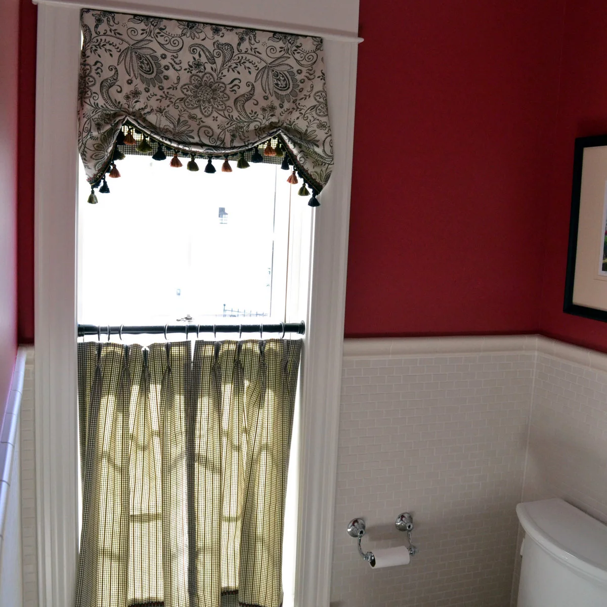 A black-and-white patterned valance with tassels sits above checked cafe curtains in a red-walled bathroom.