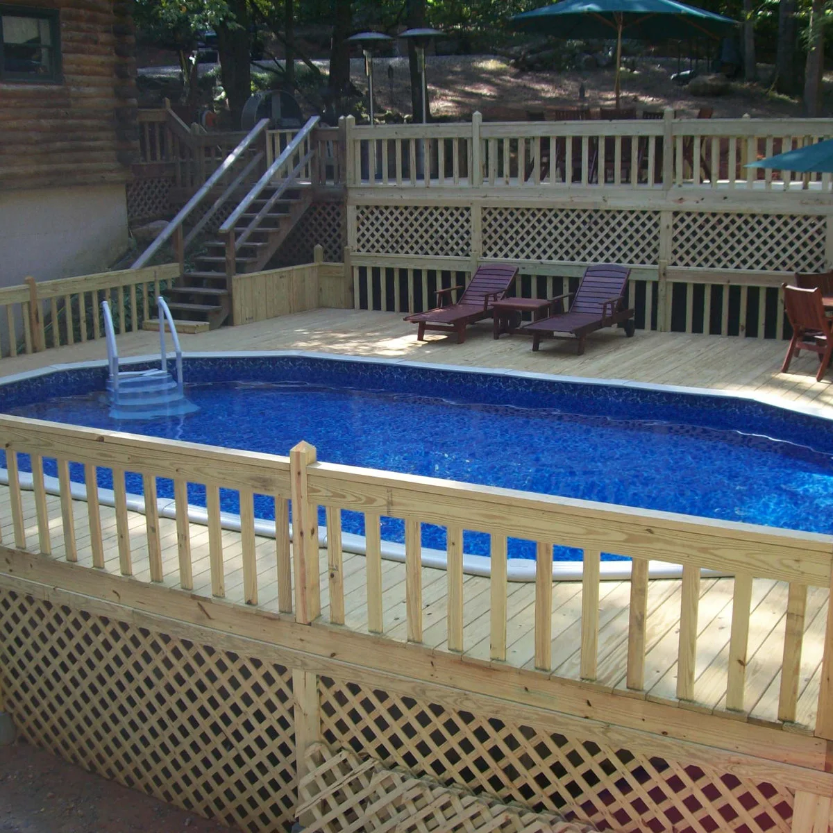 Oval above-ground pool on a lower deck with loungers and stairs connecting to a higher deck beside a wood-sided house.