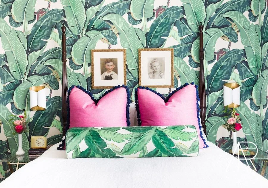Dense green banana-leaf wallpaper rises behind white bedding, two bright pink cushions and a smaller leaf-patterned bolster between wall lights
