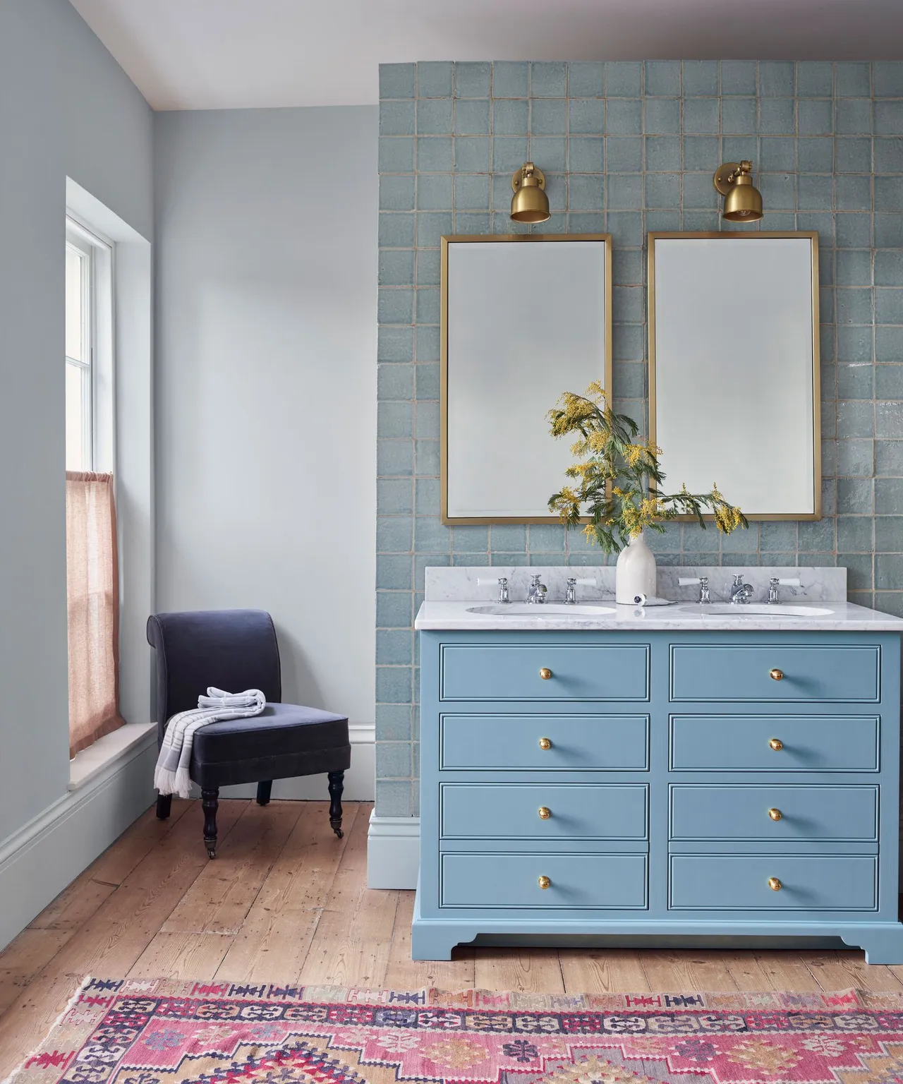 A pale blue vanity stands below simple mirrors beside a patterned rug and upholstered chair.