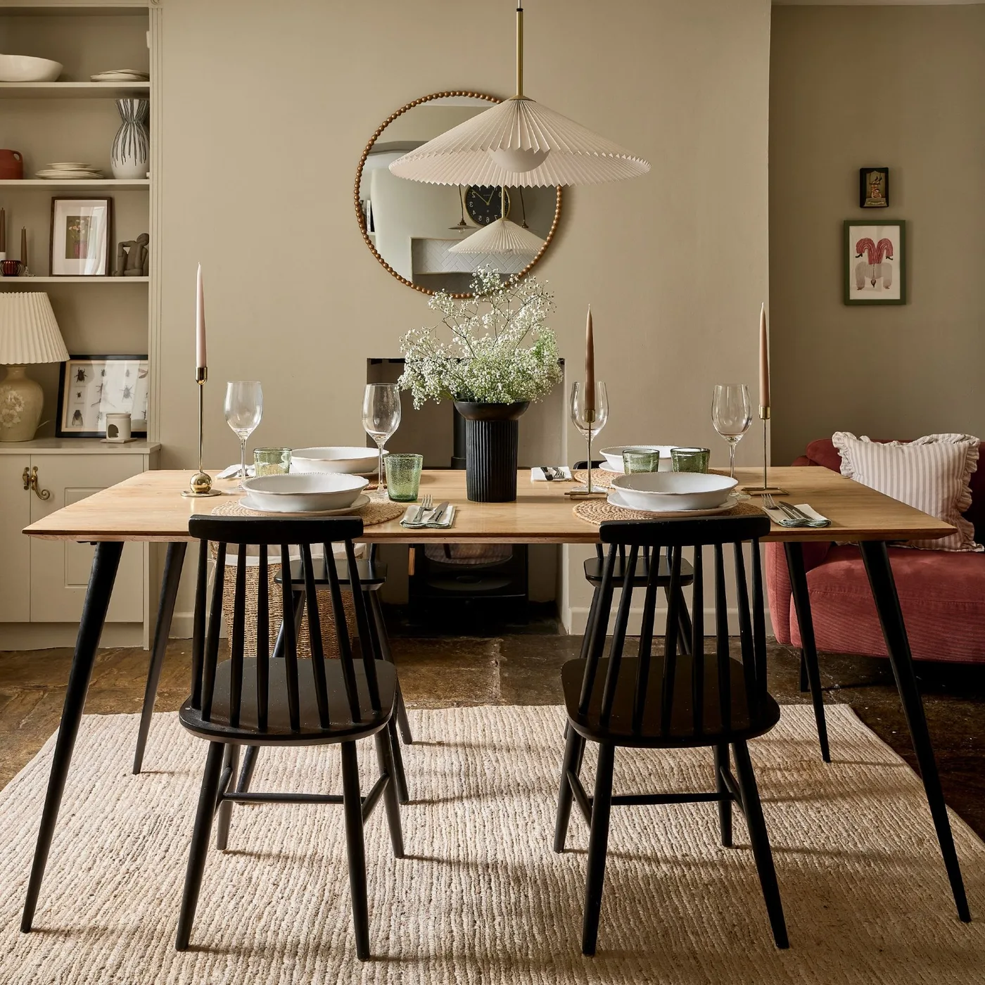 Dining room with beige walls, a wooden table, dark spindle-back chairs, round wall mirror and a pale pleated pendant.