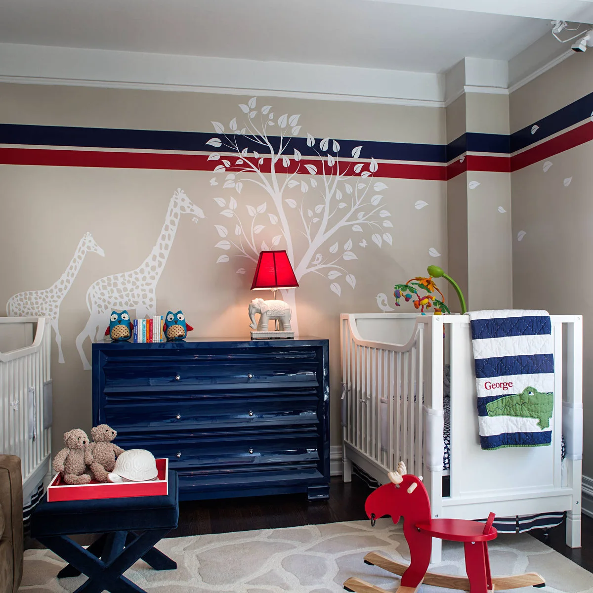 Two white cribs flanking a navy dresser beneath a white giraffe-and-tree mural and red-blue wall bands