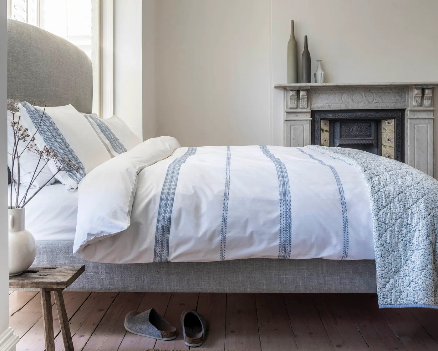 Neutral bedroom with white embroidered bedding, a blue floral throw and a pale upholstered headboard.