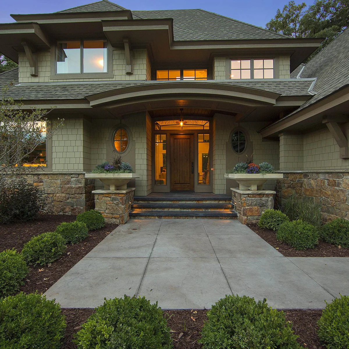 A green Craftsman house with a broad front walk, low rounded shrubs and stone entrance columns.