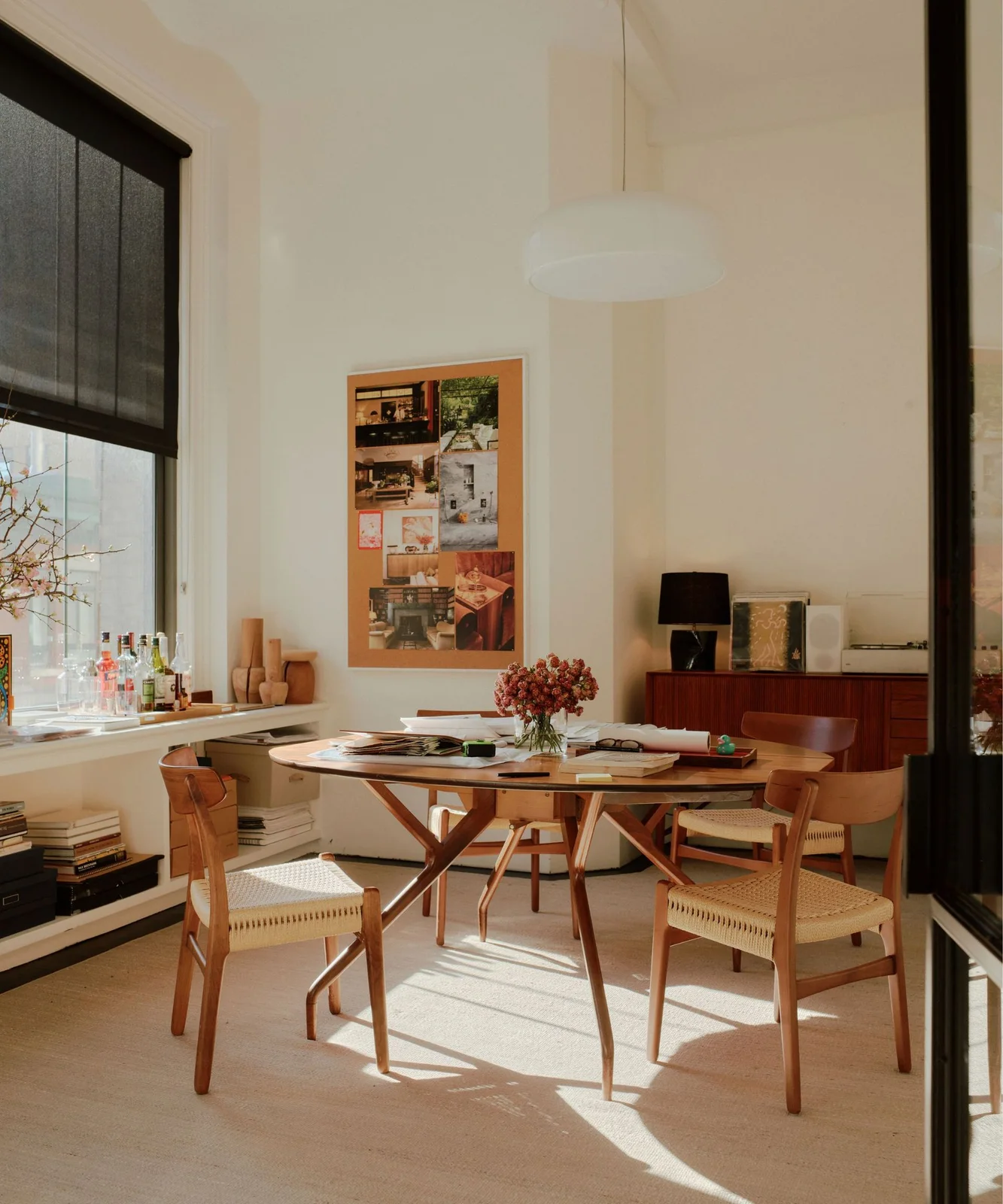 A light room has a round wood table and woven-seat wood chairs beneath a pale pendant, with papers on the table, a cork noticeboard and a low wood sideboard.