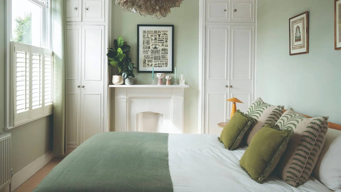 Tall white cupboards flank a white fireplace in a pale sage bedroom with a green throw, botanical pillows and a shuttered window.