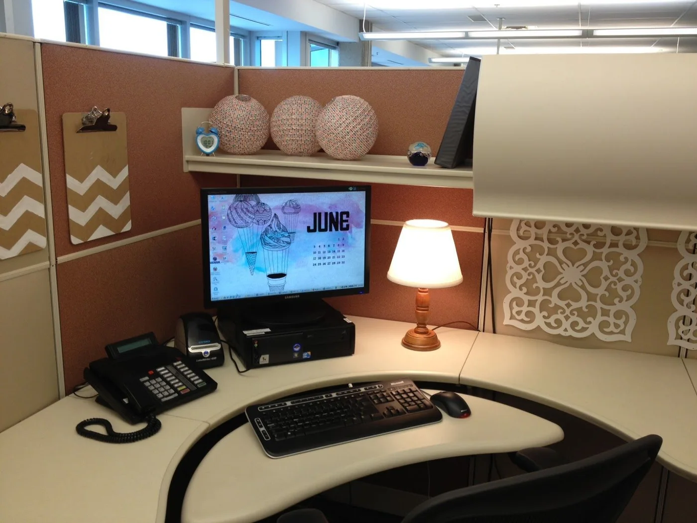 Three round paper decorations on a cubicle shelf above a monitor with chevron clipboards and a small shaded lamp.
