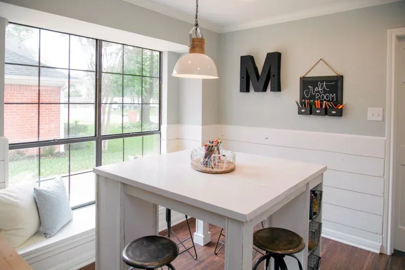 A white craft table and dark stools sit beside a broad window in a room with pale gray upper walls and white horizontal lower paneling