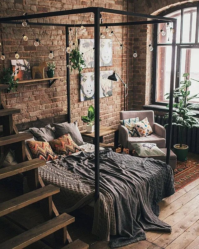 Bedroom with exposed brick, dark four-poster bed, gray textured bedding, patterned cushions, plants, and string lights.