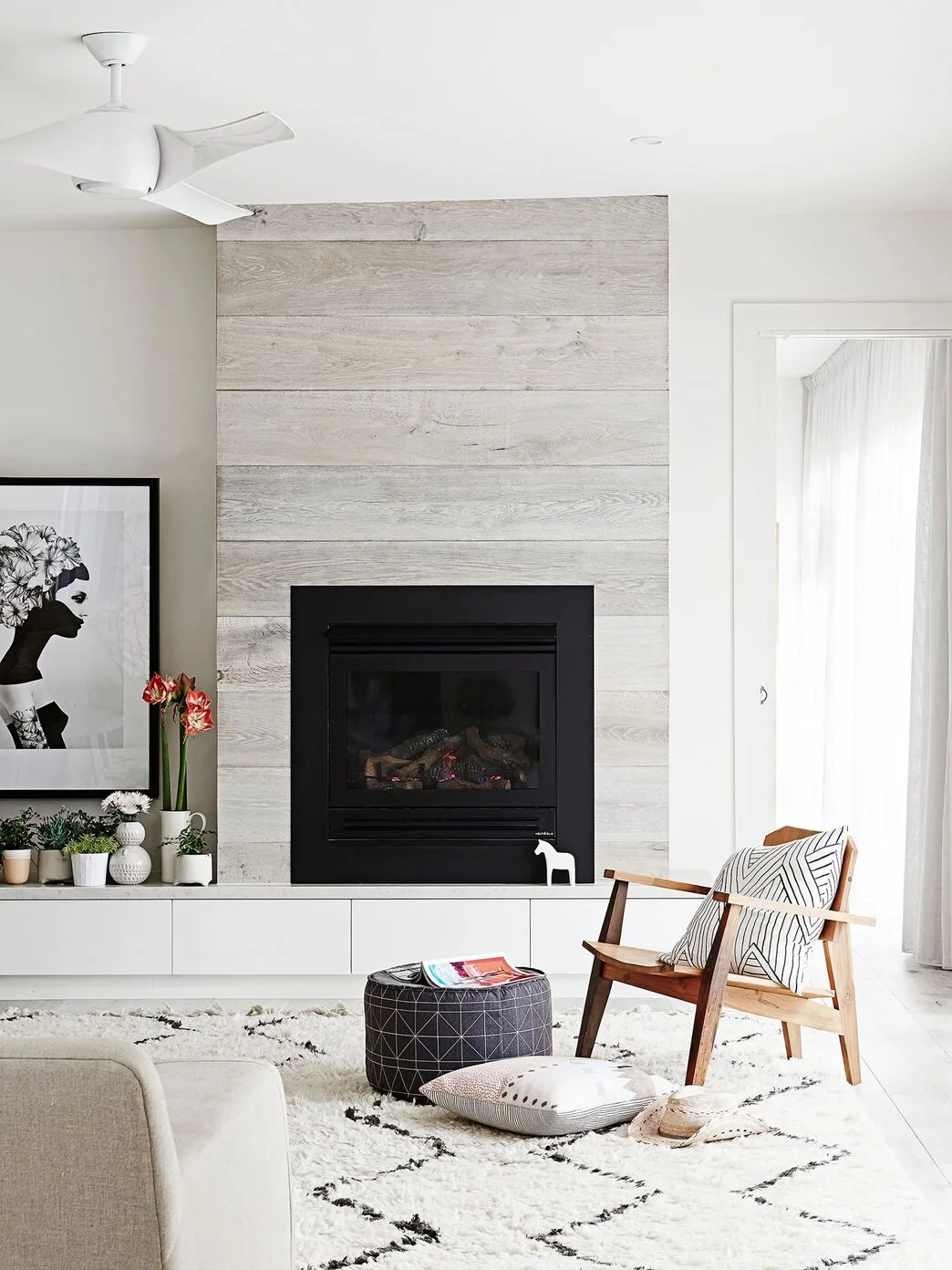 A pale wood-board chimney breast surrounds a black glazed fire above low white drawers, with a wood armchair, patterned cushions and light floor coverings.