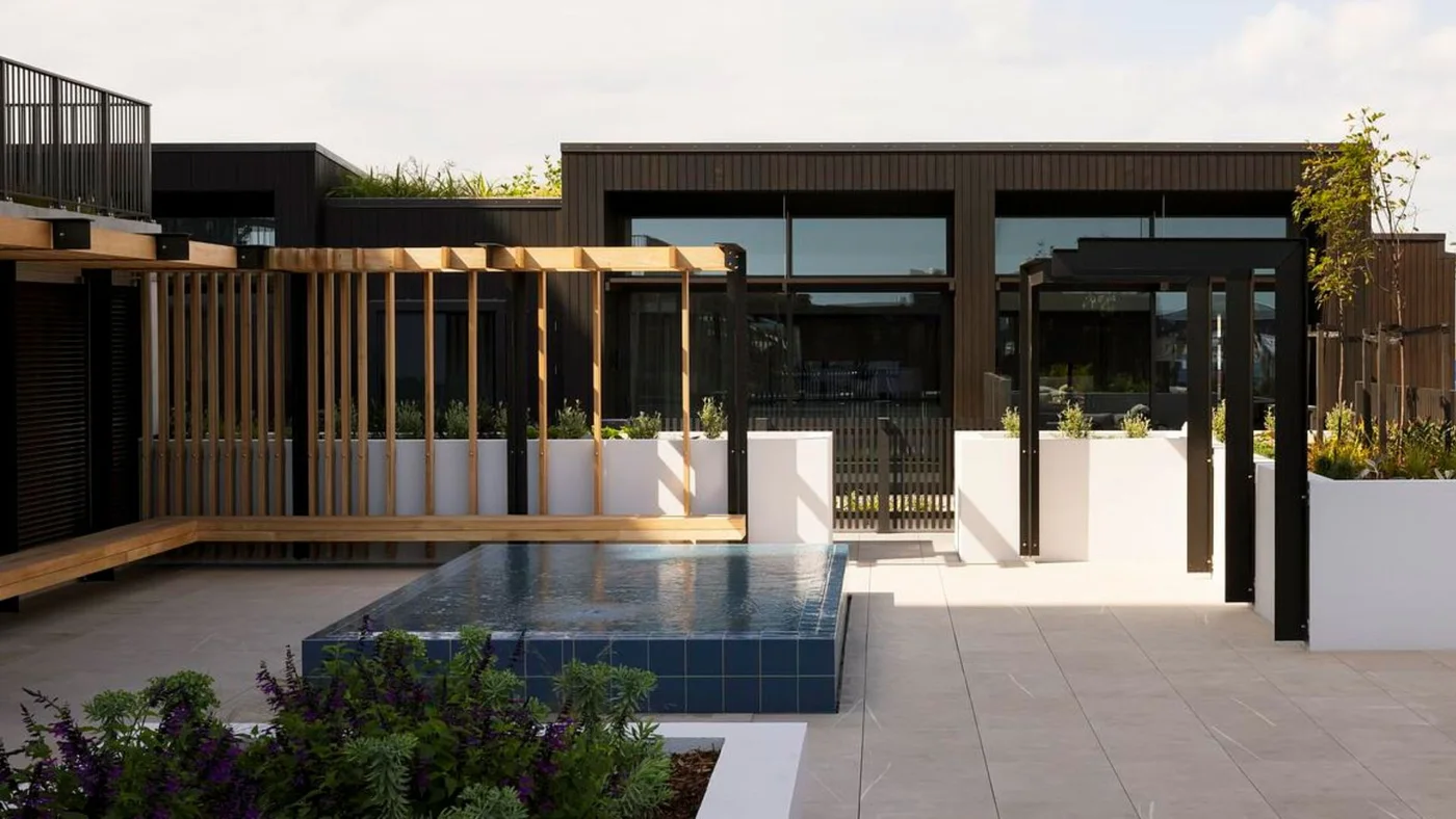A raised rectangular pool finished in dark blue square tiles sits beside pale paving, wood screens and built-in bench seating.