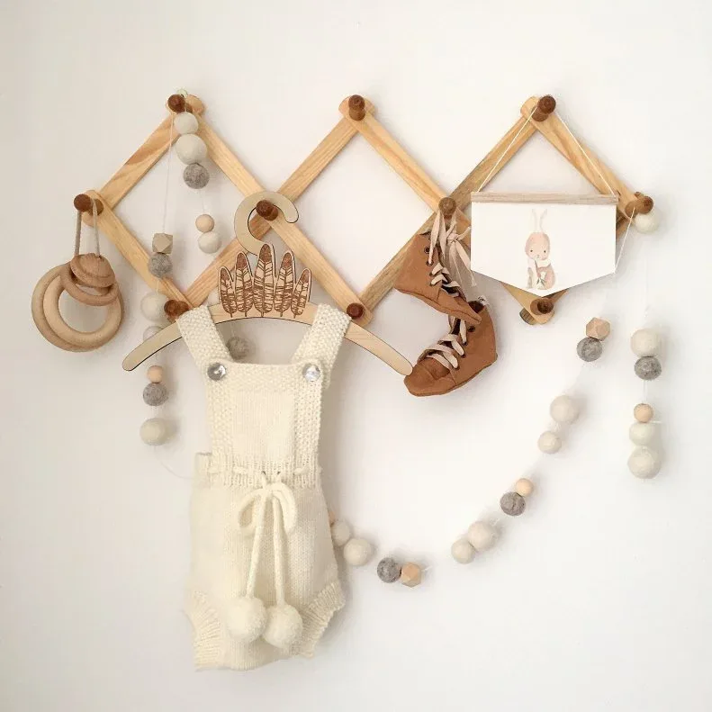 Expandable wooden peg rack on pale wall displaying small garments, artwork and hanging decorative pieces.