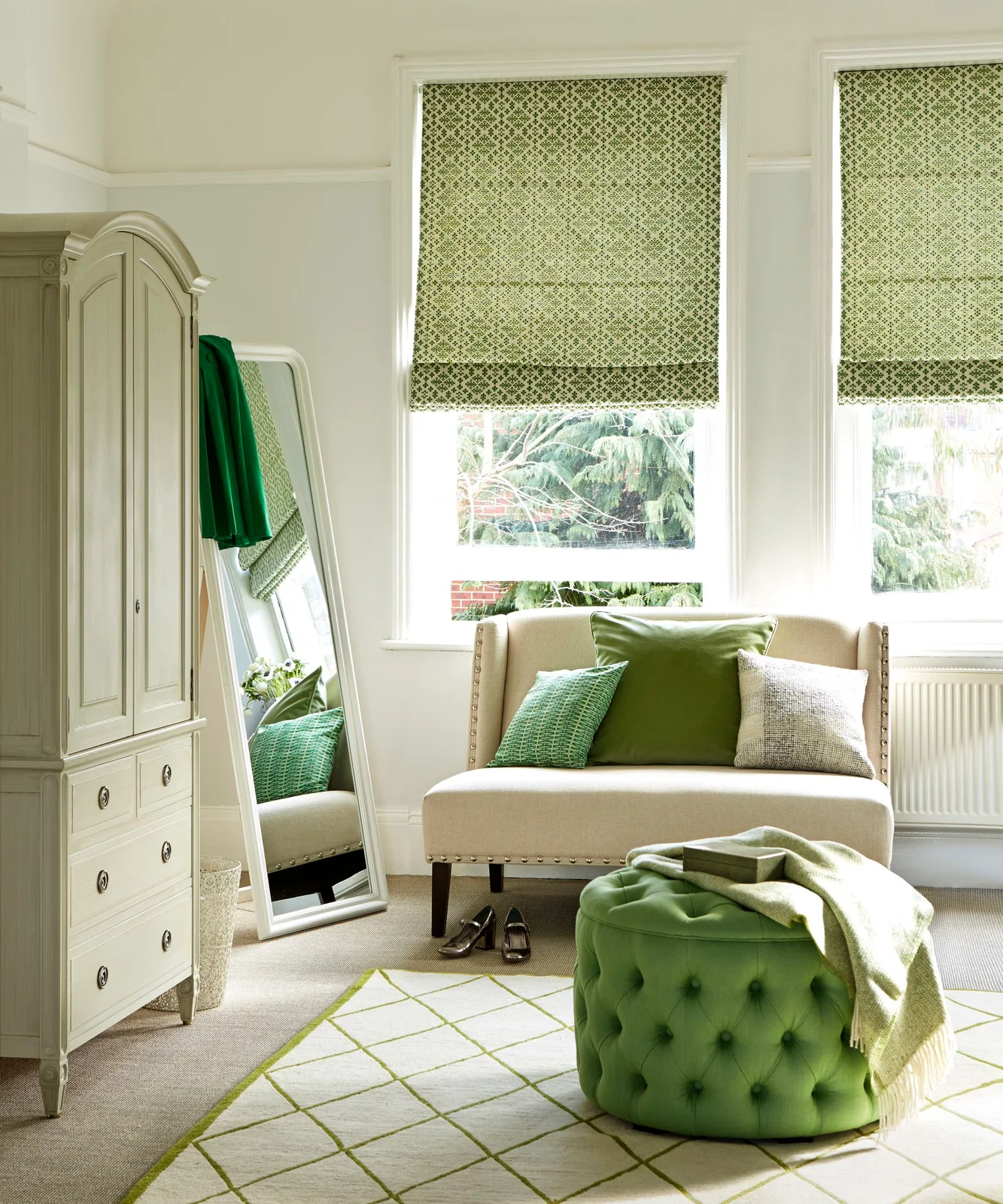A pale chair with green cushions sits near patterned green blinds, a green ottoman, pale drawers and a tall leaning mirror