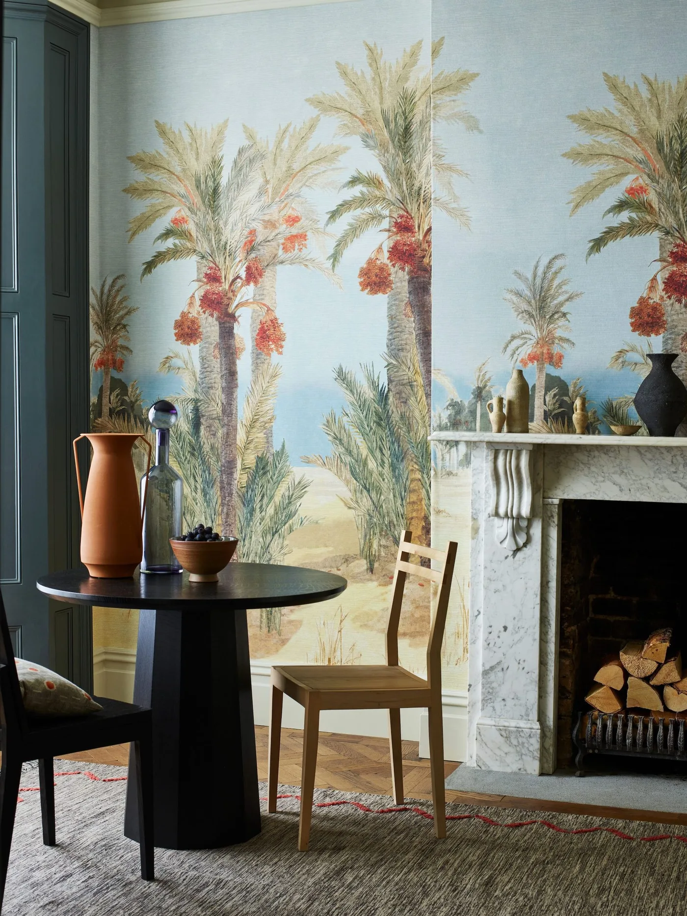 Small dining area with palm-tree wallpaper, a dark round table, contrasting chairs and a pale fireplace surround