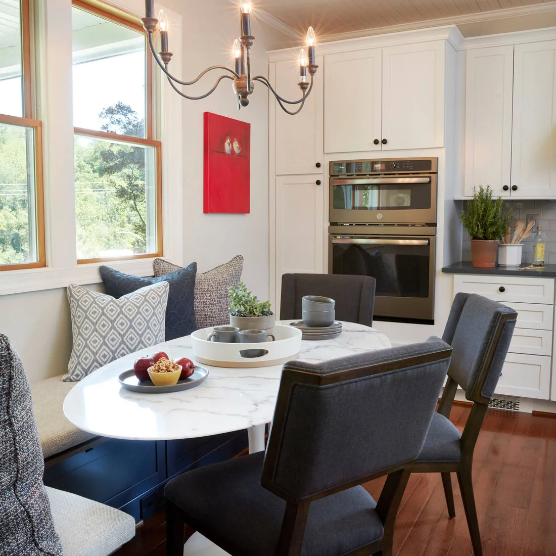 Pale oval table with gray chairs and a blue window bench beside white kitchen cabinets and ovens
