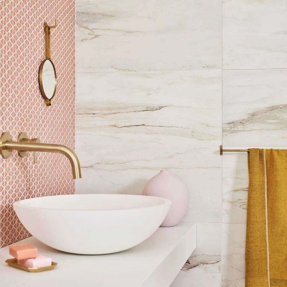 Small pink geometric tiles form a sink backsplash beside pale veined wall tiles and gold-colored fittings