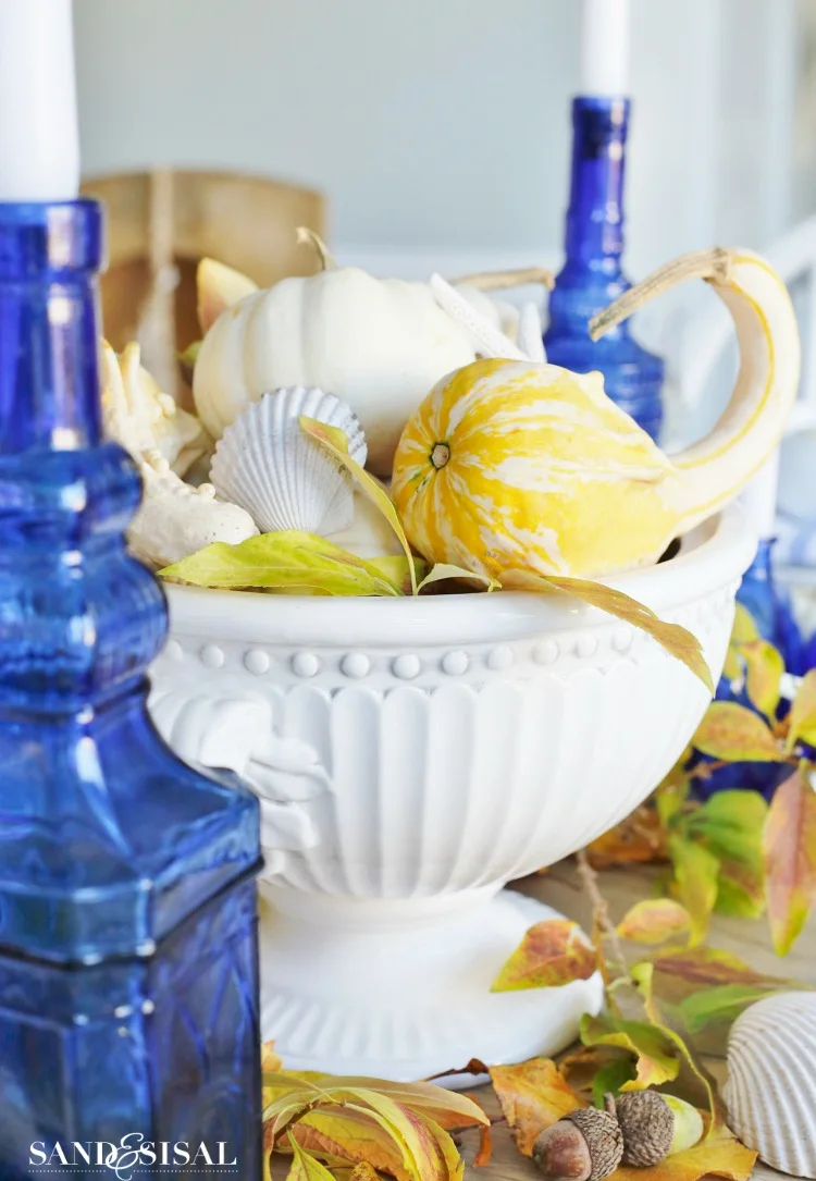 White pumpkins and striped yellow gourds fill a white footed bowl surrounded by leaves, shells, a starfish shape and blue candle bottles