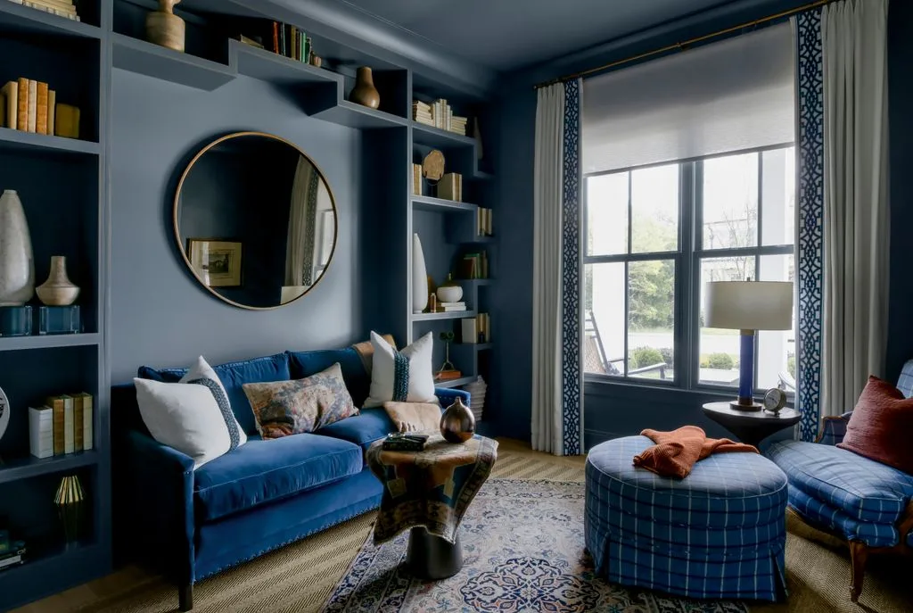 Dark blue bedroom sitting area with built-in shelves, blue seating, plaid ottoman, patterned rug, and round mirror.