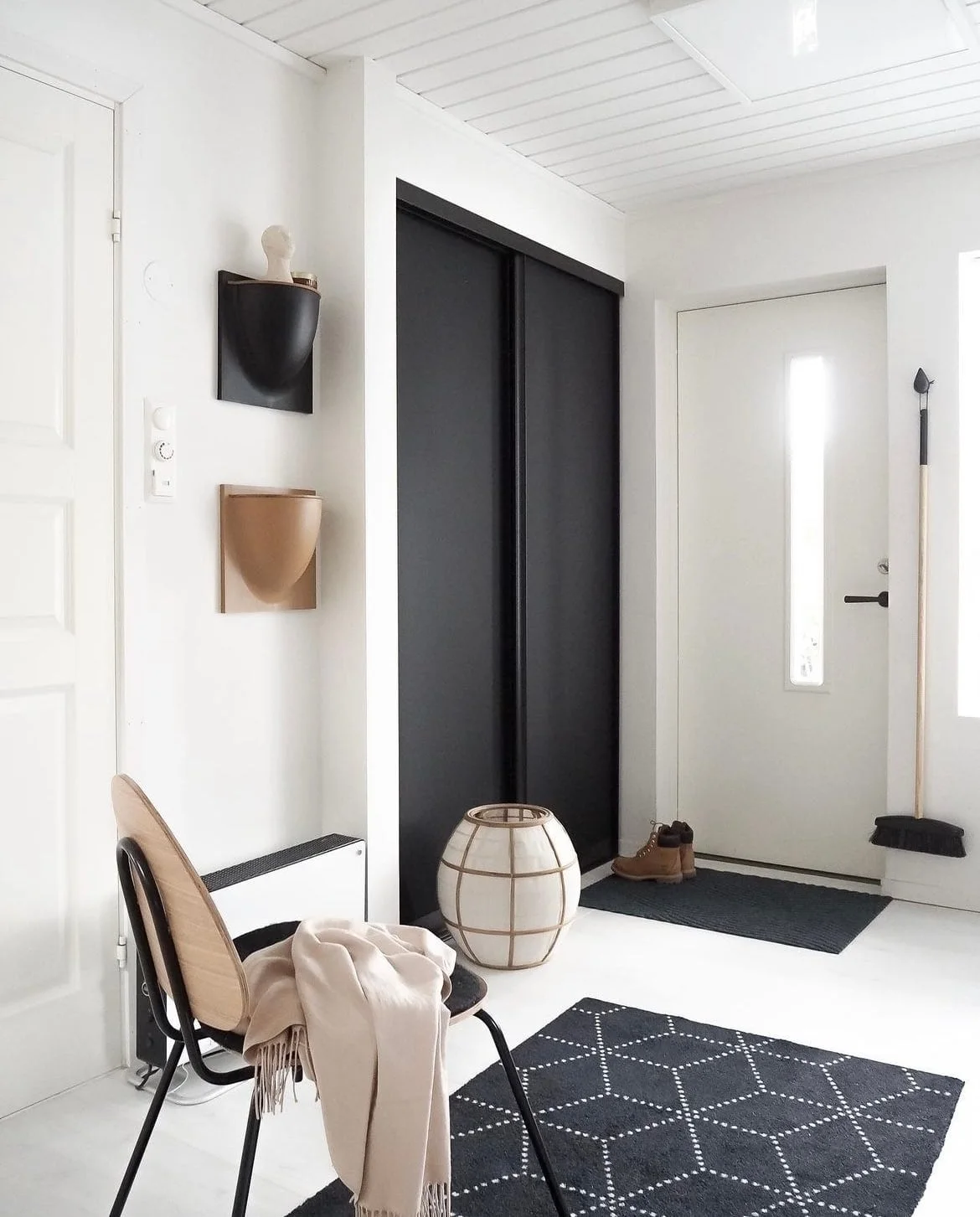 A wooden chair with a black frame sits beside a tall black storage door, small wall pockets, a pale rounded lamp and dark patterned rug