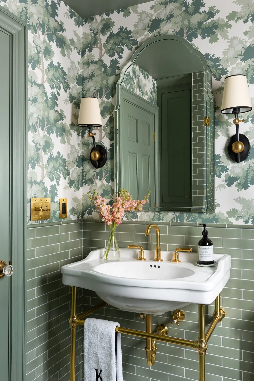 Sage subway-tile wainscoting sits beneath tree-patterned wallpaper around a brass console basin, shaped mirror and shaded wall lights.