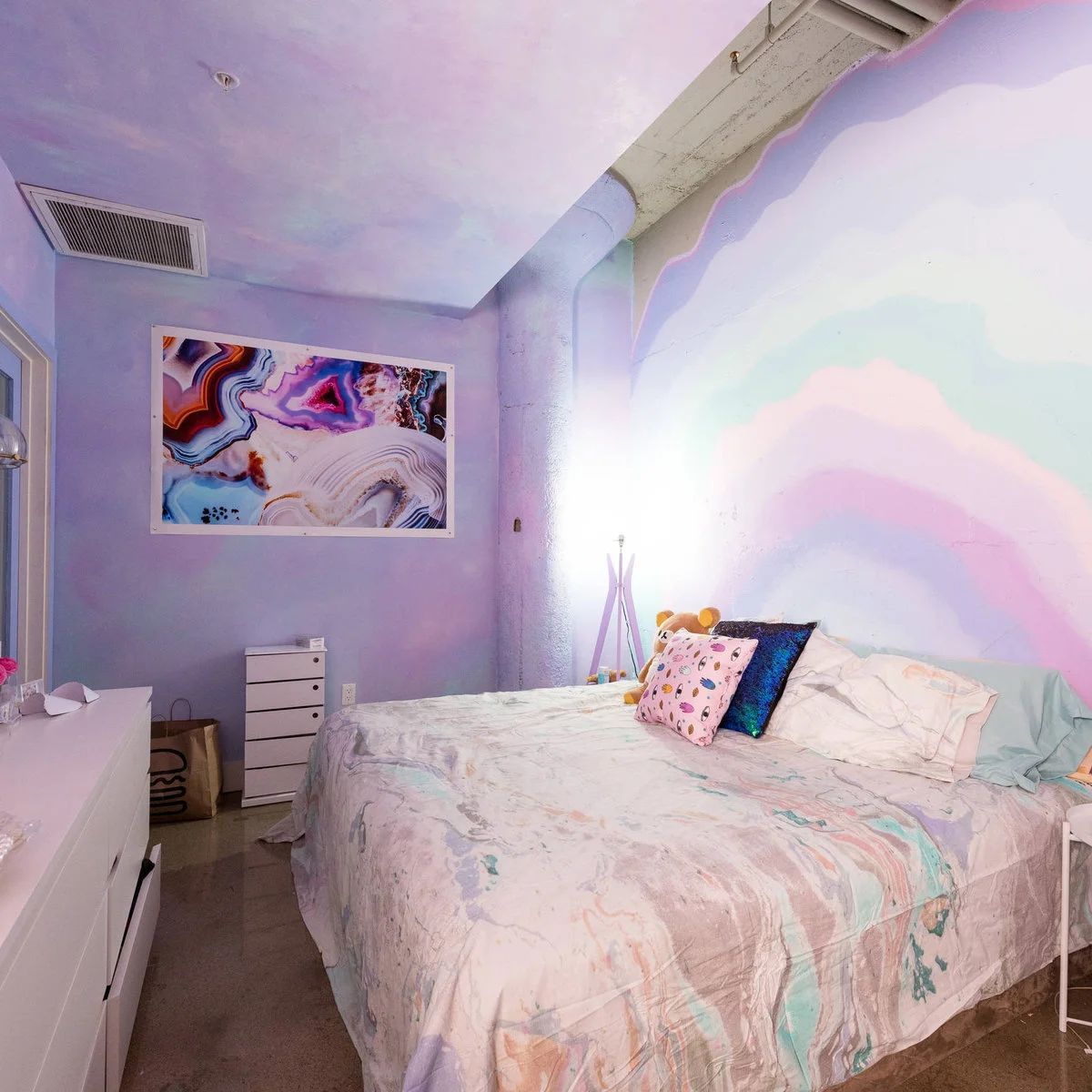 Lavender bedroom with a broad pastel rainbow wall design, white furniture and marbled-pattern bedding