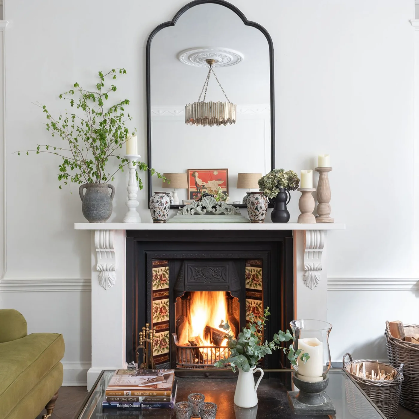 Floral tiles flank a dark fireplace insert beneath a pale carved mantel and tall shaped mirror.