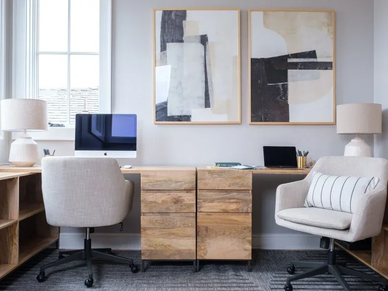 Two work surfaces meet at wood drawer cabinets beneath abstract pictures, with a rolling desk chair, an upholstered chair and pale lamps.