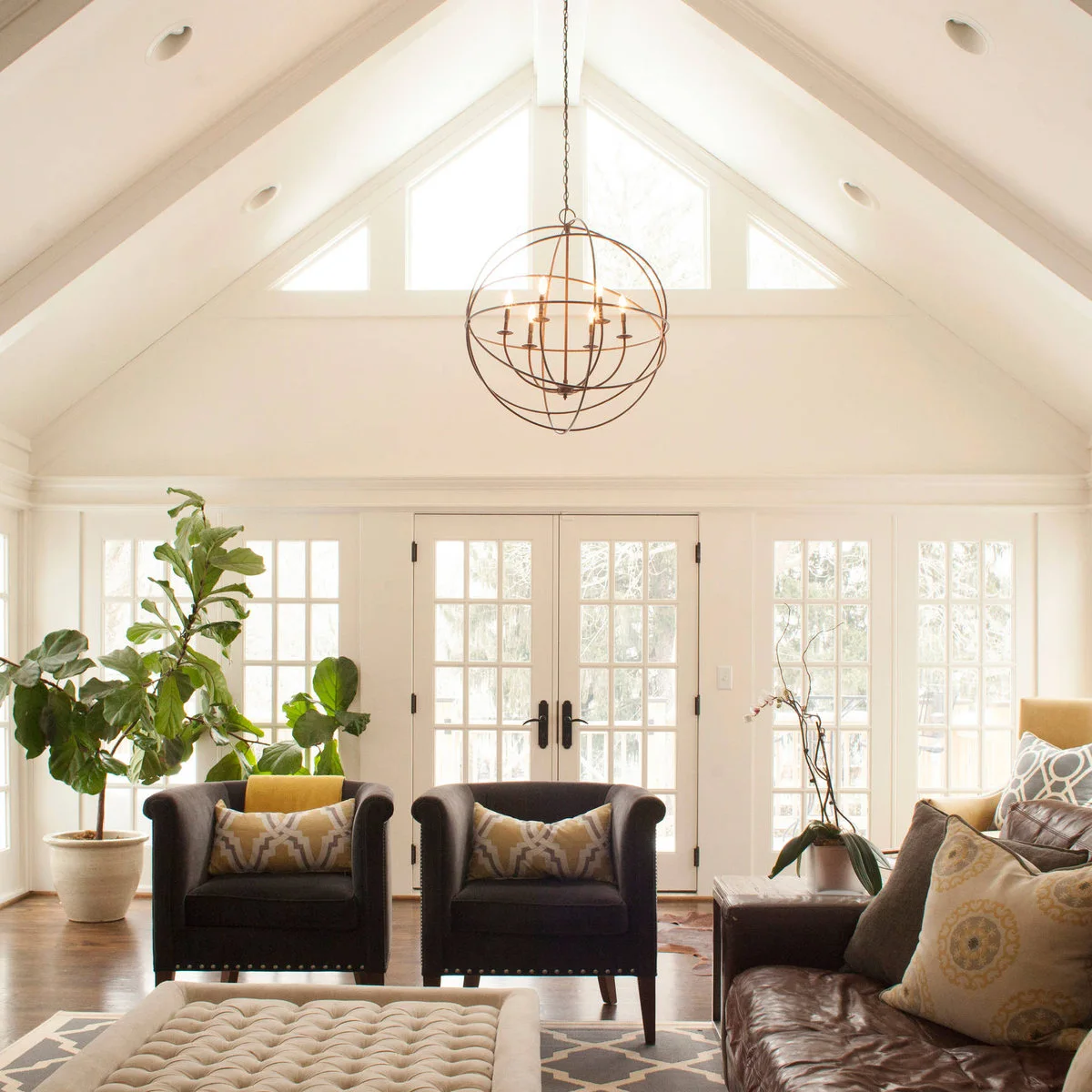 Two dark upholstered chairs and a leather sofa in a pale sunroom with a high ceiling, glazed doors and large plants.