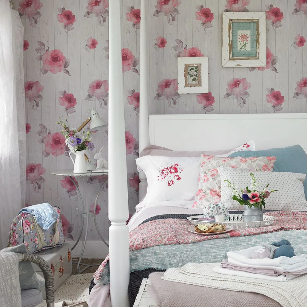 Pink roses printed over pale plank-effect wallpaper sit behind a white four-poster bed with floral cushions.