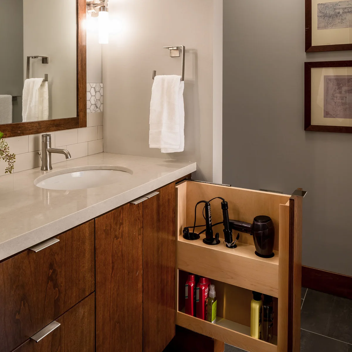 A white towel hangs from an angular open-ended holder beside a wood-framed mirror above a pale counter and wood vanity.