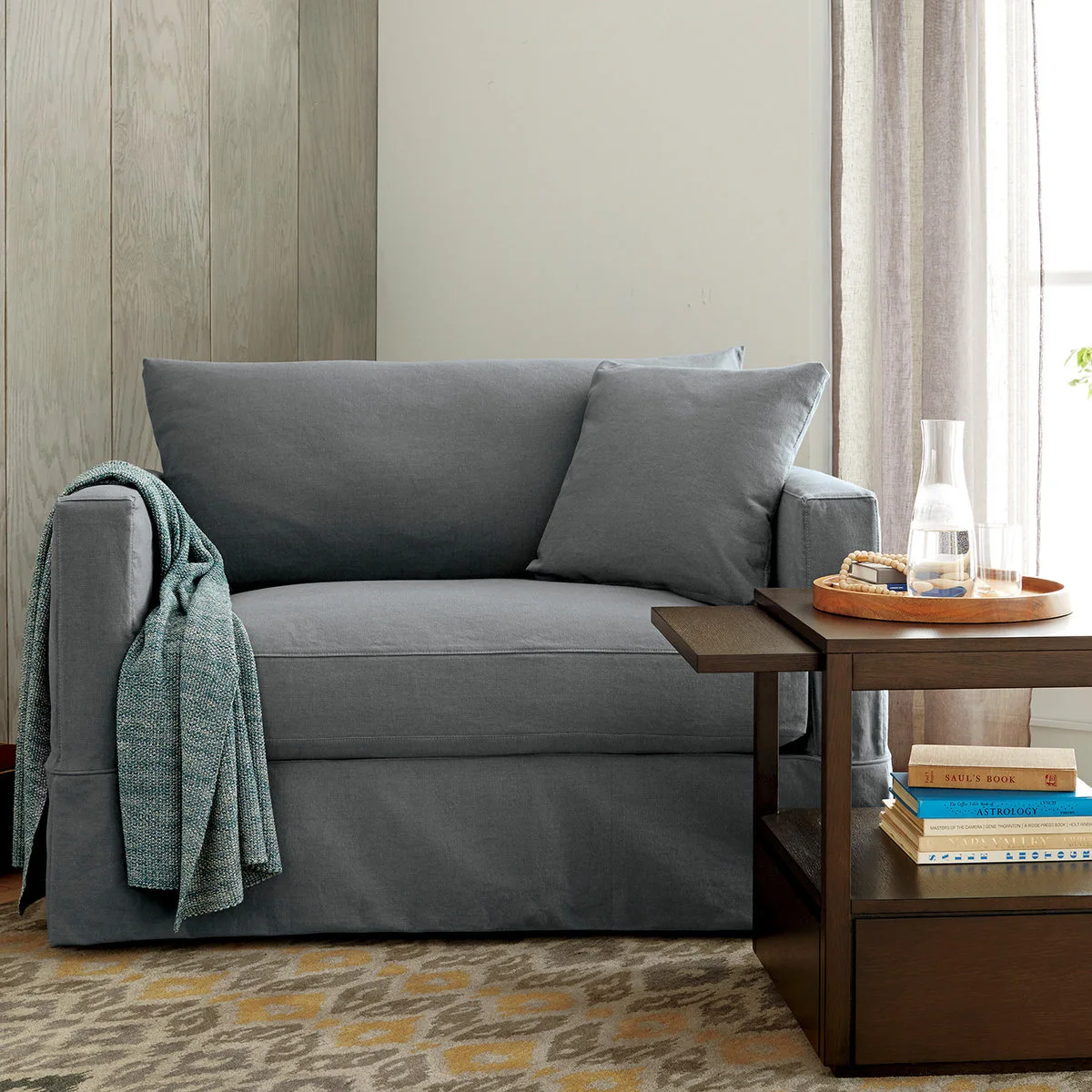 Compact gray slipcovered sleeper chair beside a wood table with an extended side ledge