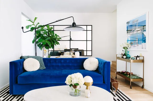 A vivid blue sofa with round pale cushions sits in a bright room beneath a black-framed mirror, beside a plant and wheeled drinks cart.