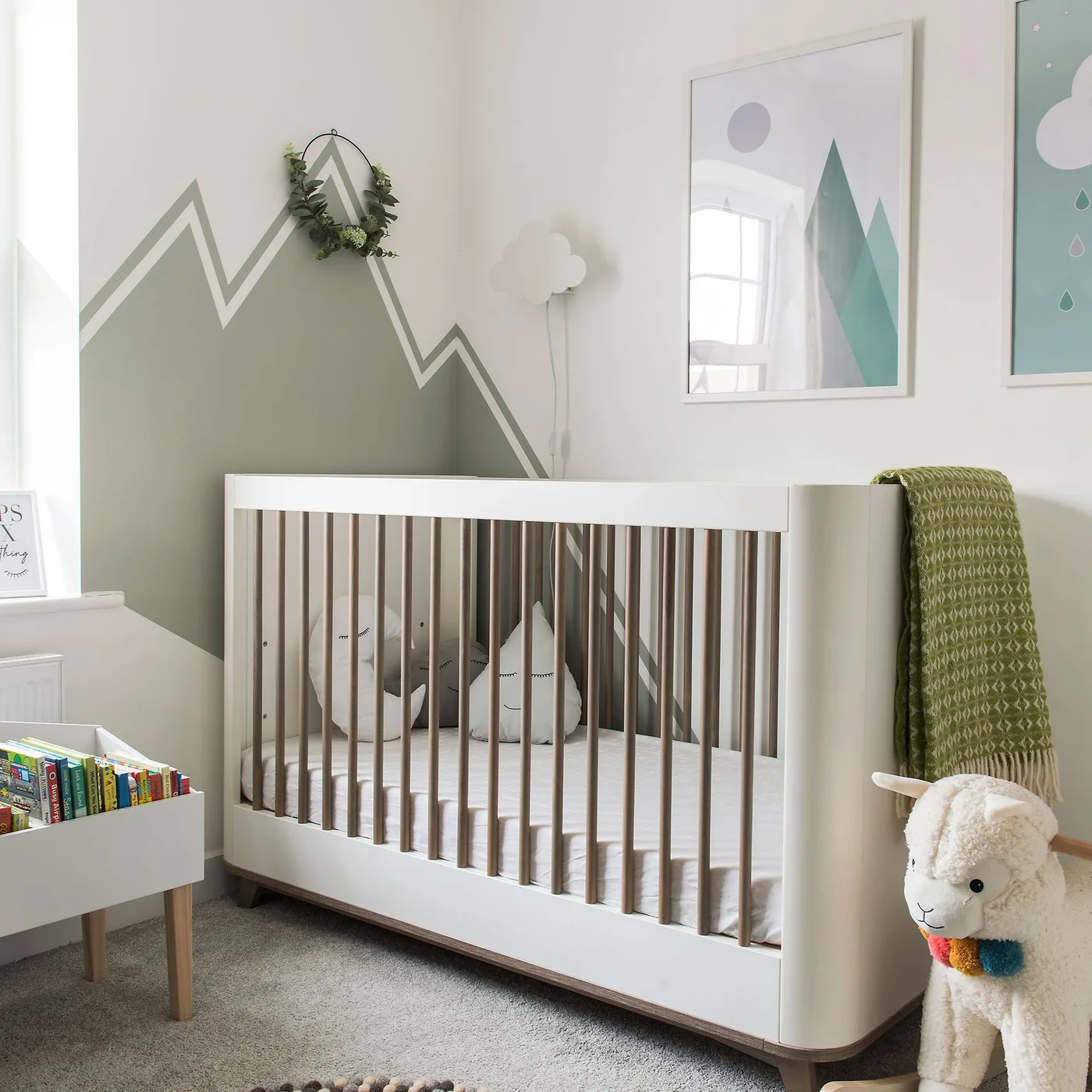 A white crib with wooden slats stands beneath a muted green geometric mural and three framed mountain pictures