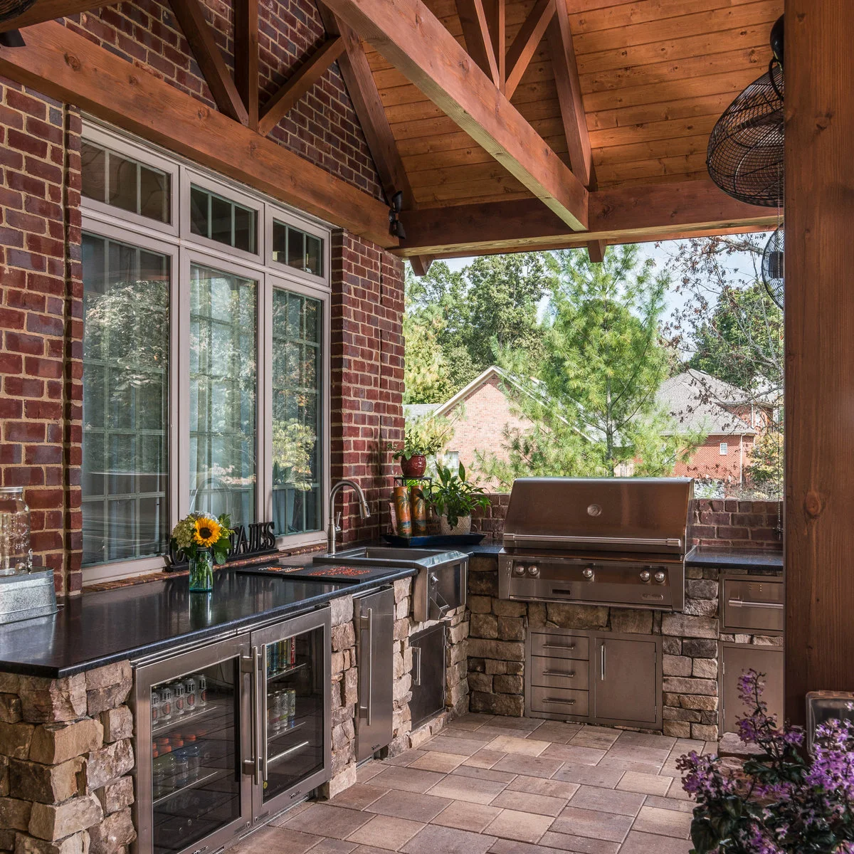 Covered outdoor kitchen with stone-faced bases, dark counters, metal storage doors, sink and beverage refrigerator