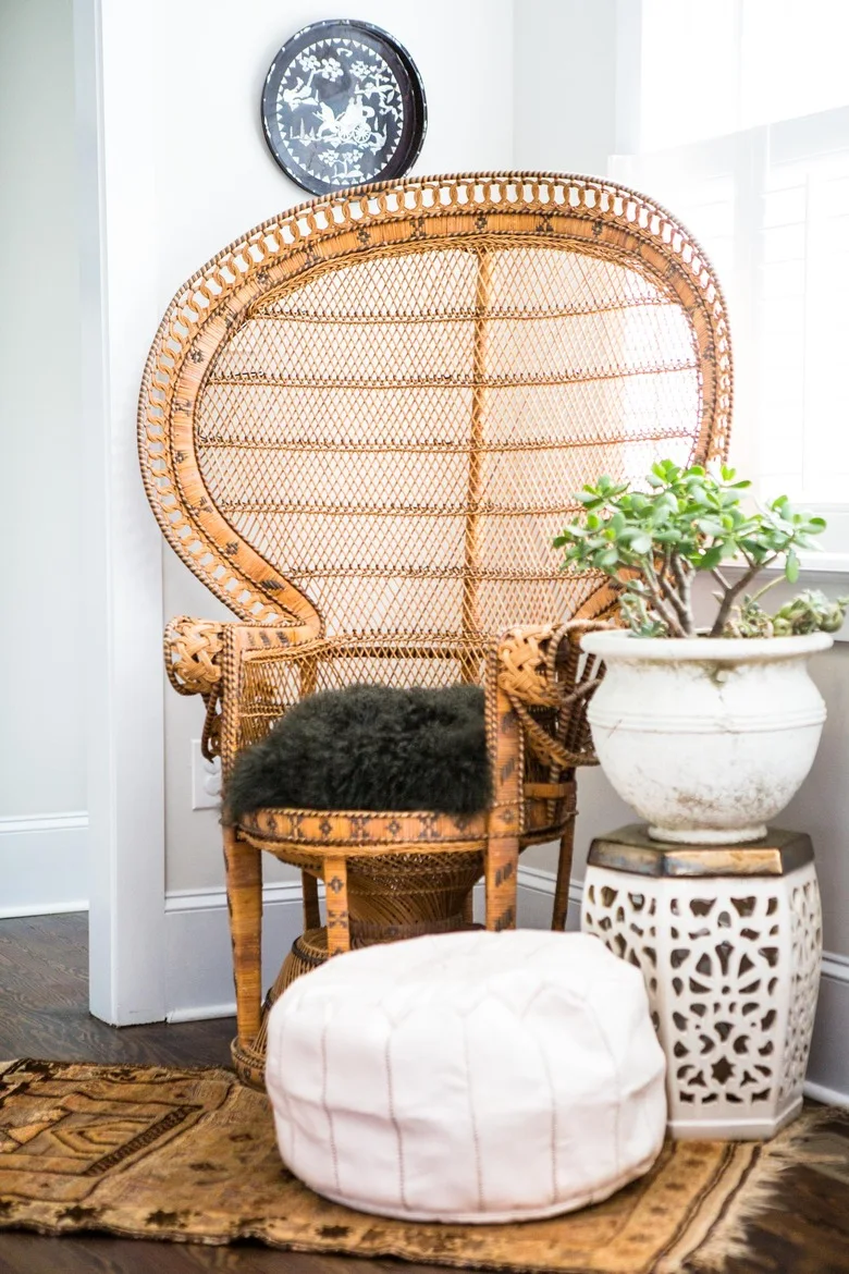 A tall woven peacock chair with a dark seat cushion sits beside a pale round floor cushion, a small pierced side table and a potted plant.
