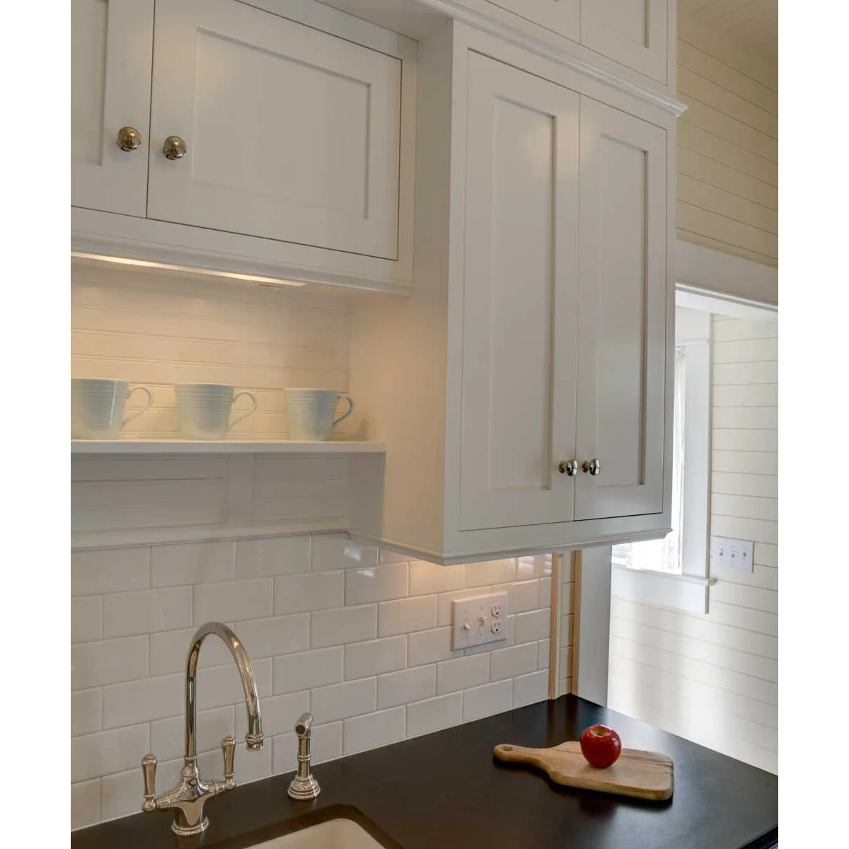 A narrow shelf with three pale cups between white upper cabinets above a kitchen sink and tiled wall.