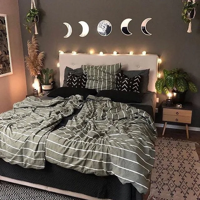 Dark bedroom wall with moon-phase mirrors, pale headboard, green checked bedding, black patterned cushions, plants, and string lights.