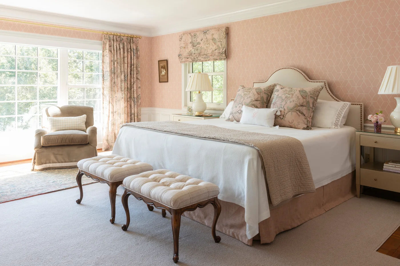 Pale pink geometric wallpaper surrounds a cream bed, floral curtains and two upholstered footstools.