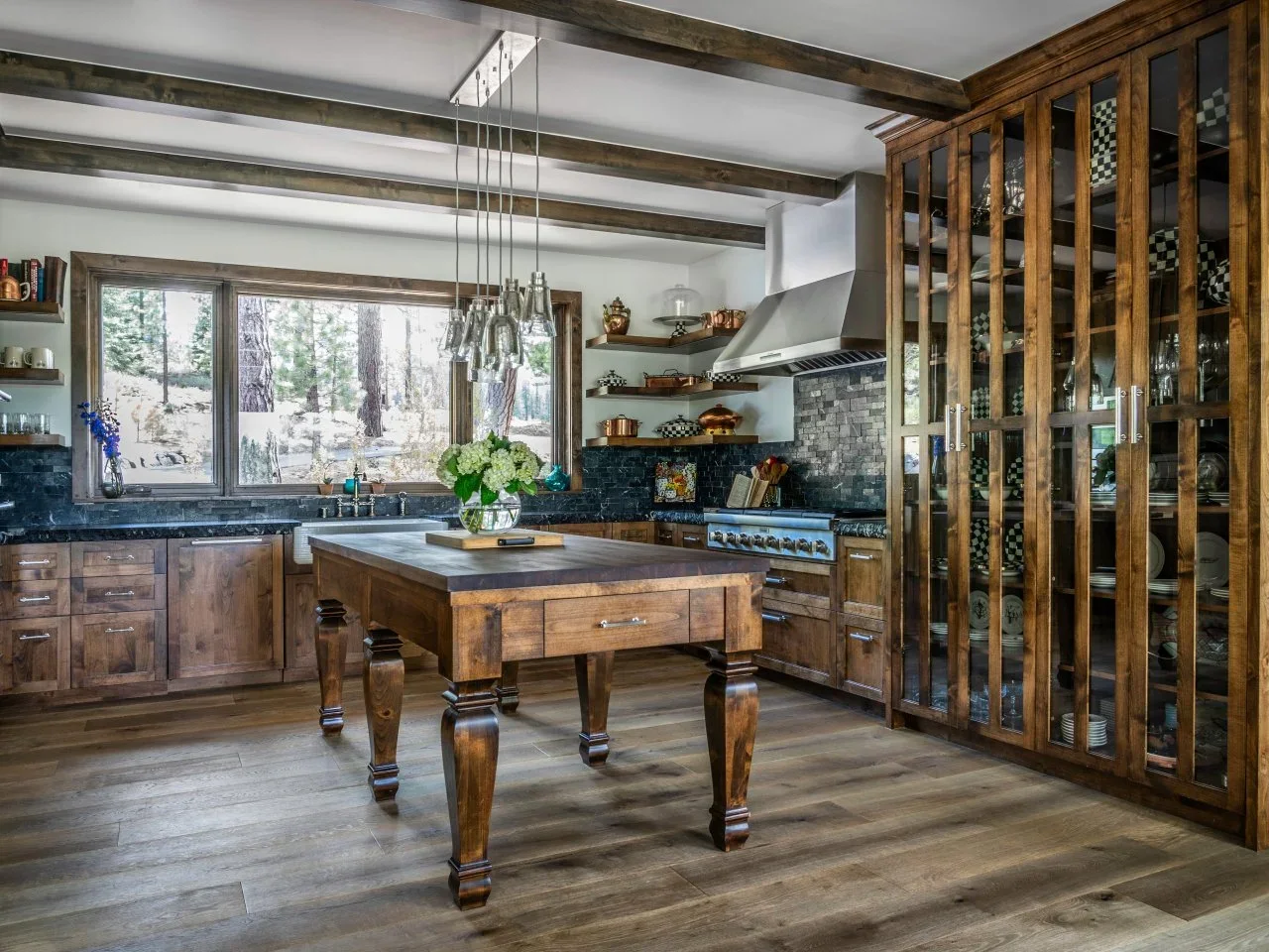 A timber island with turned legs stands among dark wood cabinets, glass-front storage and exposed ceiling beams.