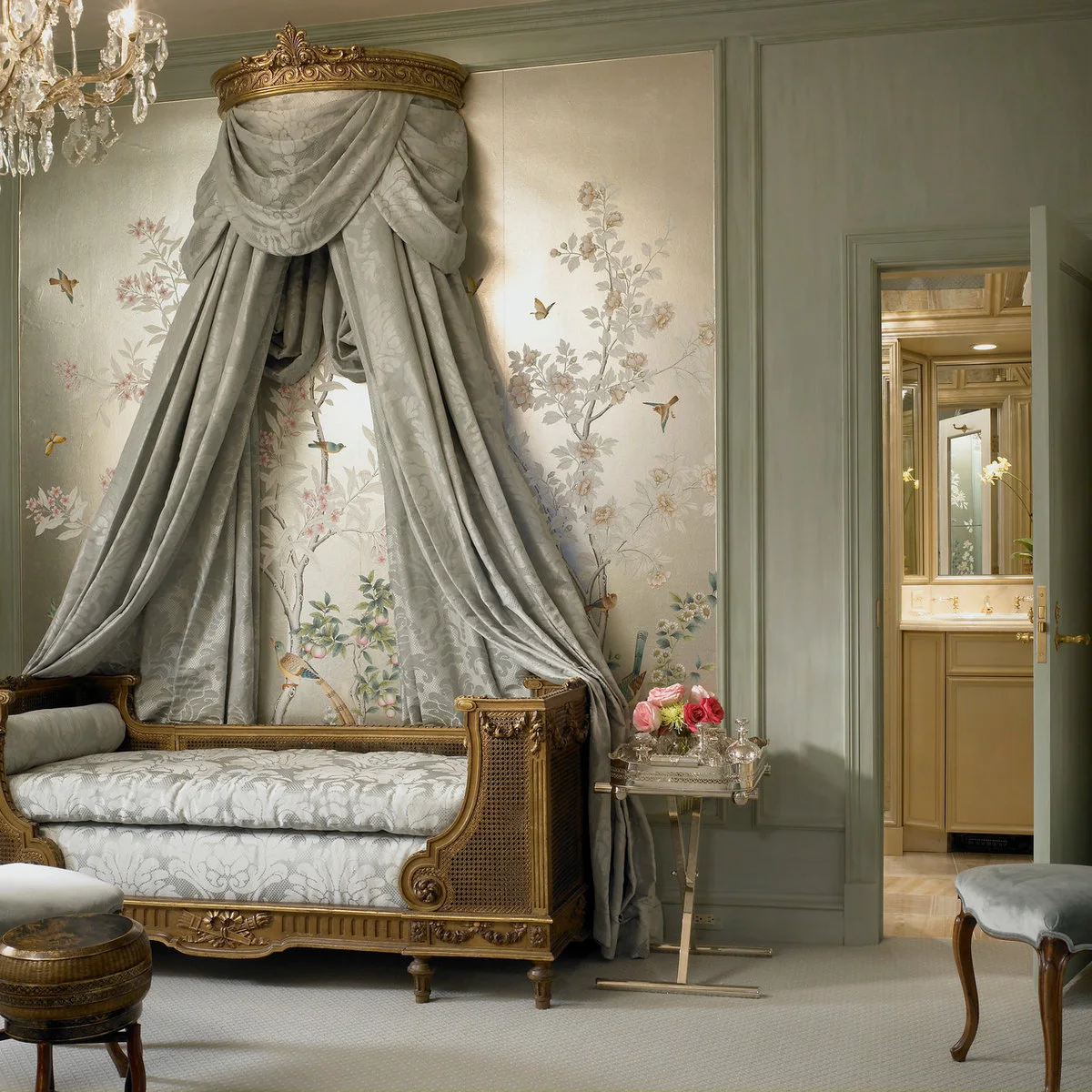 An ornate gold-colored daybed with pale patterned upholstery sits below a draped canopy against a botanical wall.