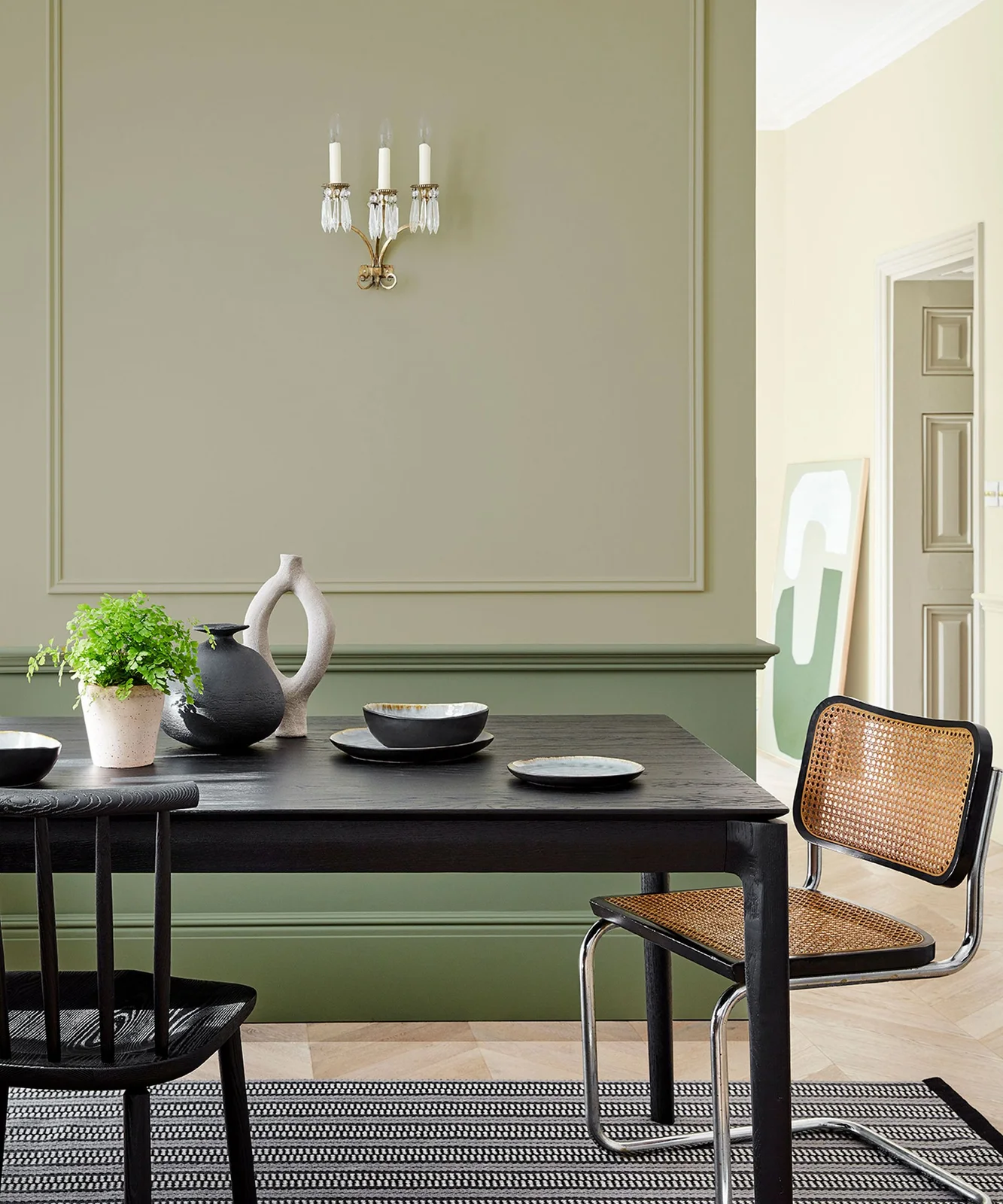Two muted green wall shades and narrow trim frame a dark dining table beside a woven-seat chair and simple pale vessels