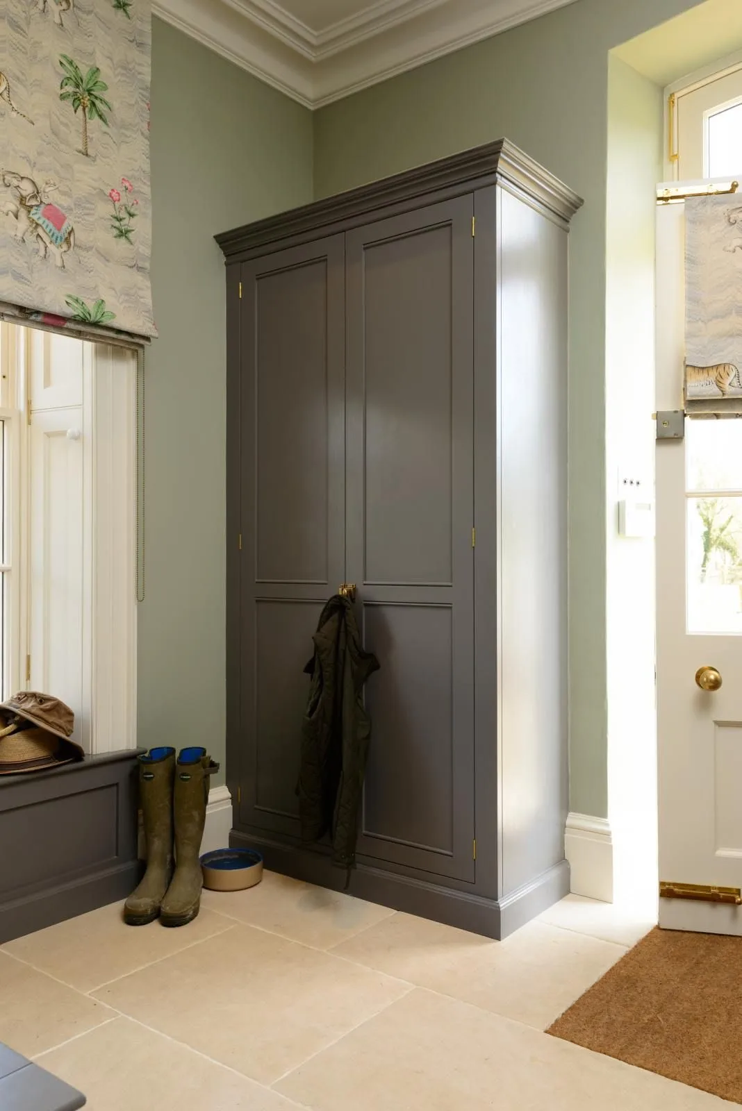 Mudroom corner with tall gray wardrobe, pale green walls, patterned blind, glazed door and boots.