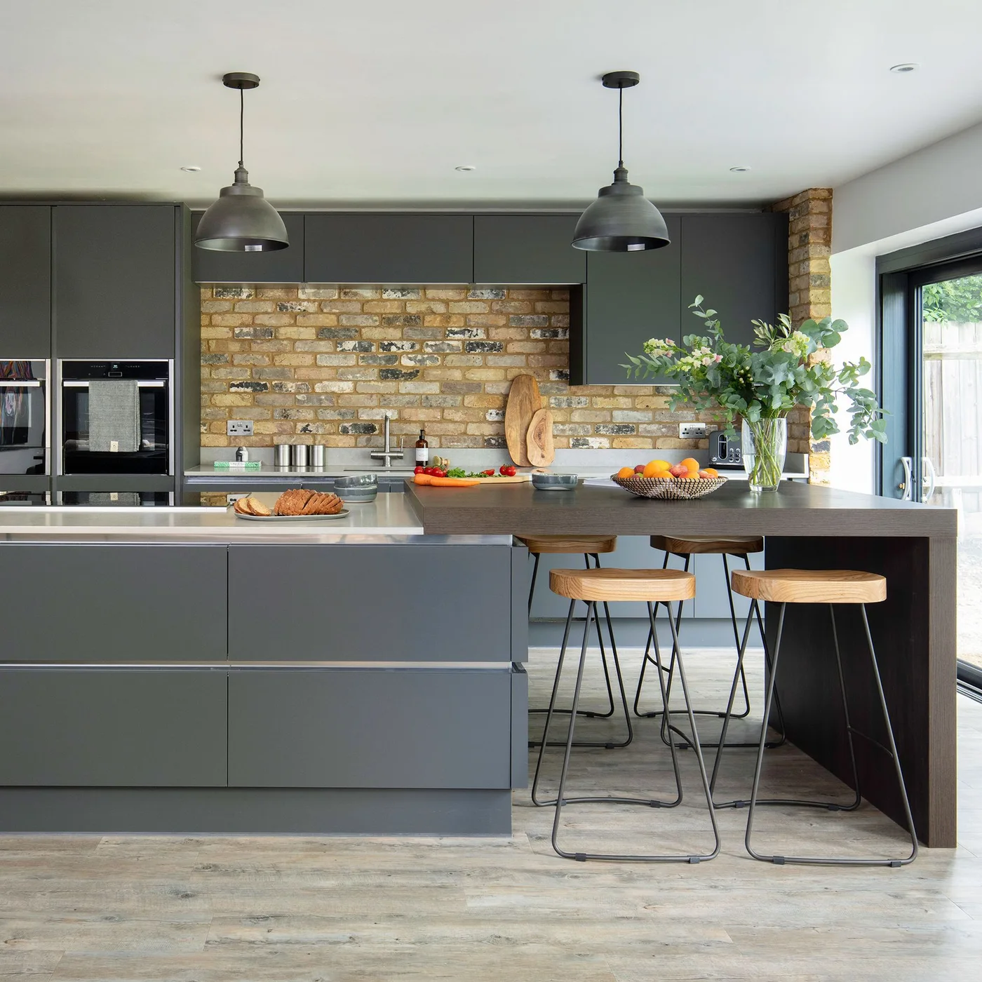 Flat gray kitchen cupboards and drawers sit beside a warm brick backsplash and a narrow seating end with two wood-topped stools