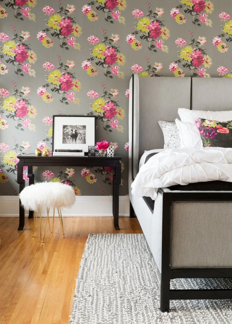 Pink and yellow flowers repeat on gray wallpaper beside a tall gray headboard and black bedside desk.