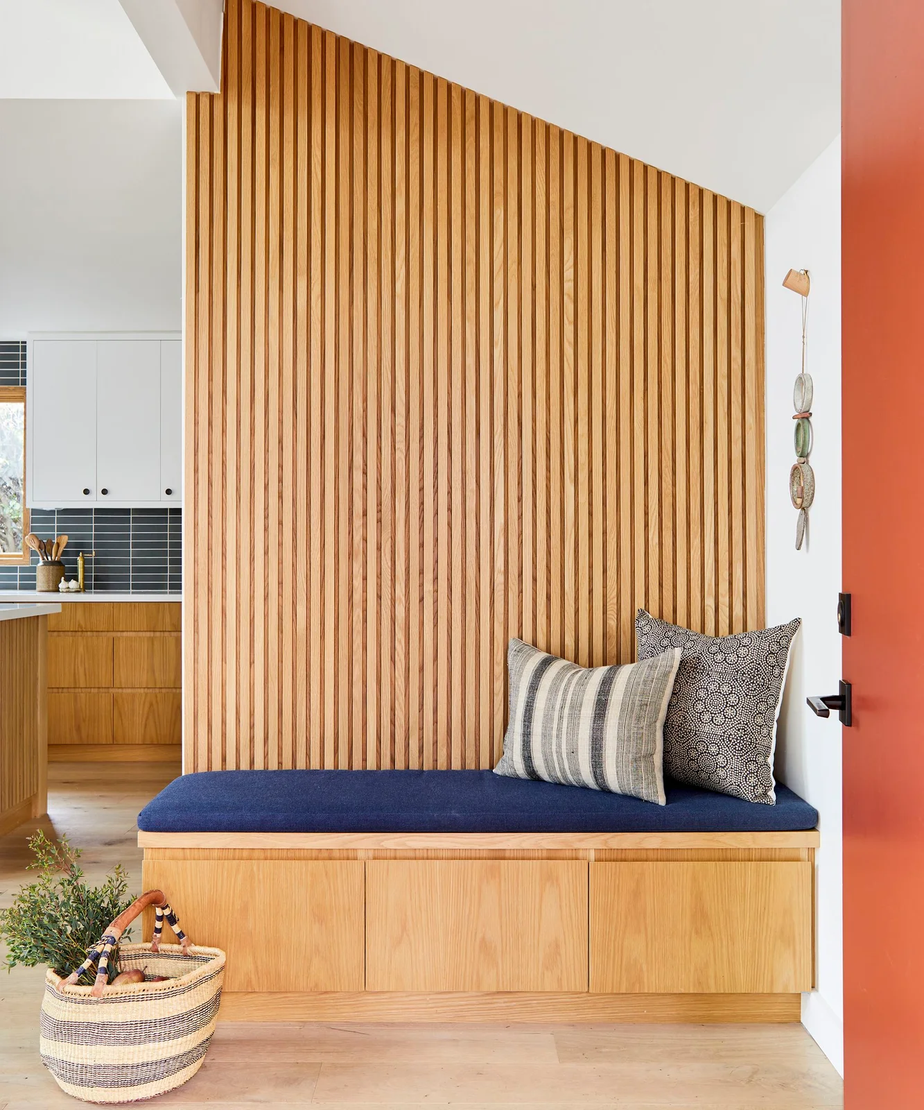A wood bench with flat closed fronts and a blue cushion sits against a tall slatted wood wall beside an orange entry door.