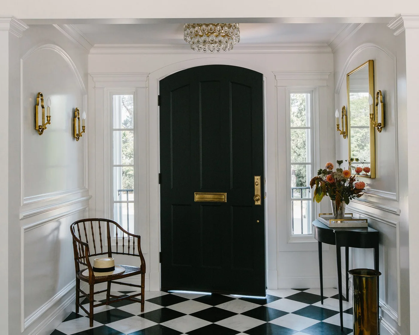 A dark arched front door sits between white walls and sidelights above black-and-white checked flooring beside a dark table and warm-colored wall fittings