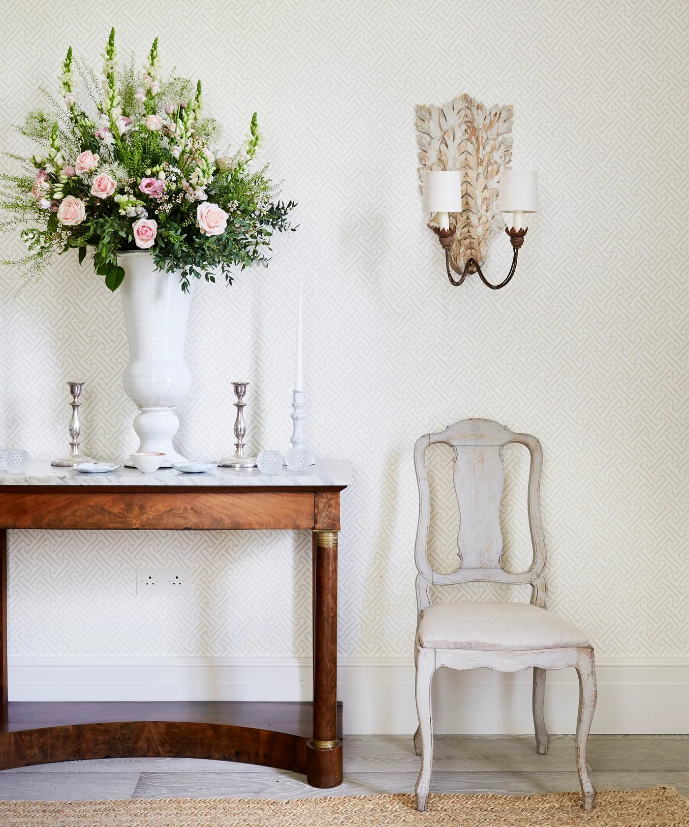 Wood console with tall white vase of pink flowers, small candlesticks and pale chair beneath a decorative wall sconce