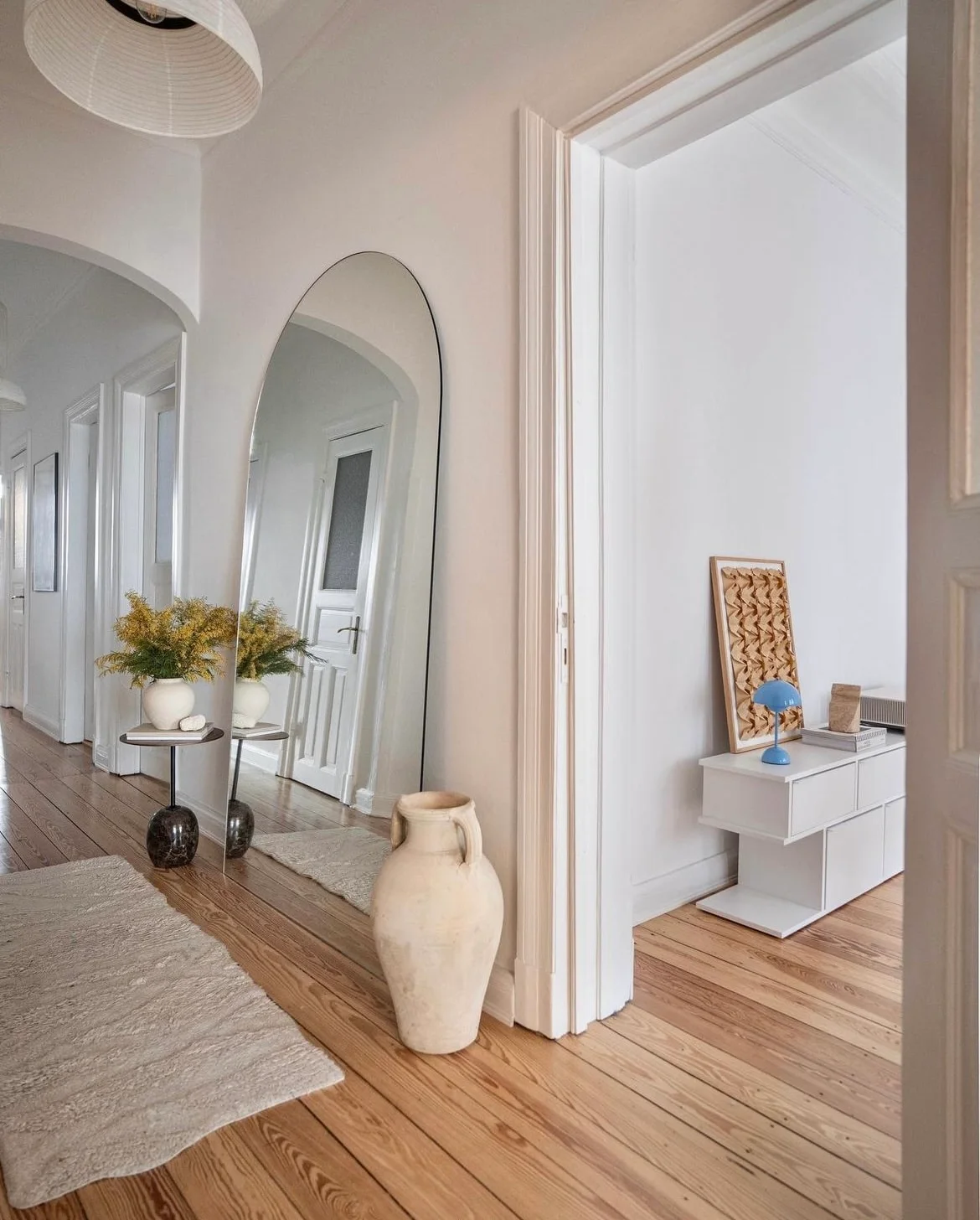 A tall rounded mirror leans beside a small round table, pale vessel and wooden floor beneath a paper-style pendant in a white hallway