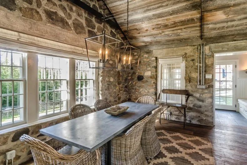 A dark dining table and woven armchairs stand in a stone-walled room with timber ceiling boards and two lantern pendants.