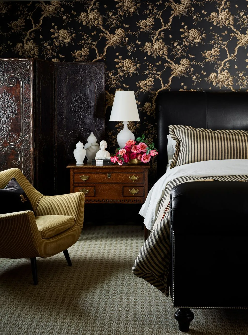 Dark floral bedroom wallpaper with a carved folding screen, striped bedding, wooden bedside chest, white lamp, and ochre chair.